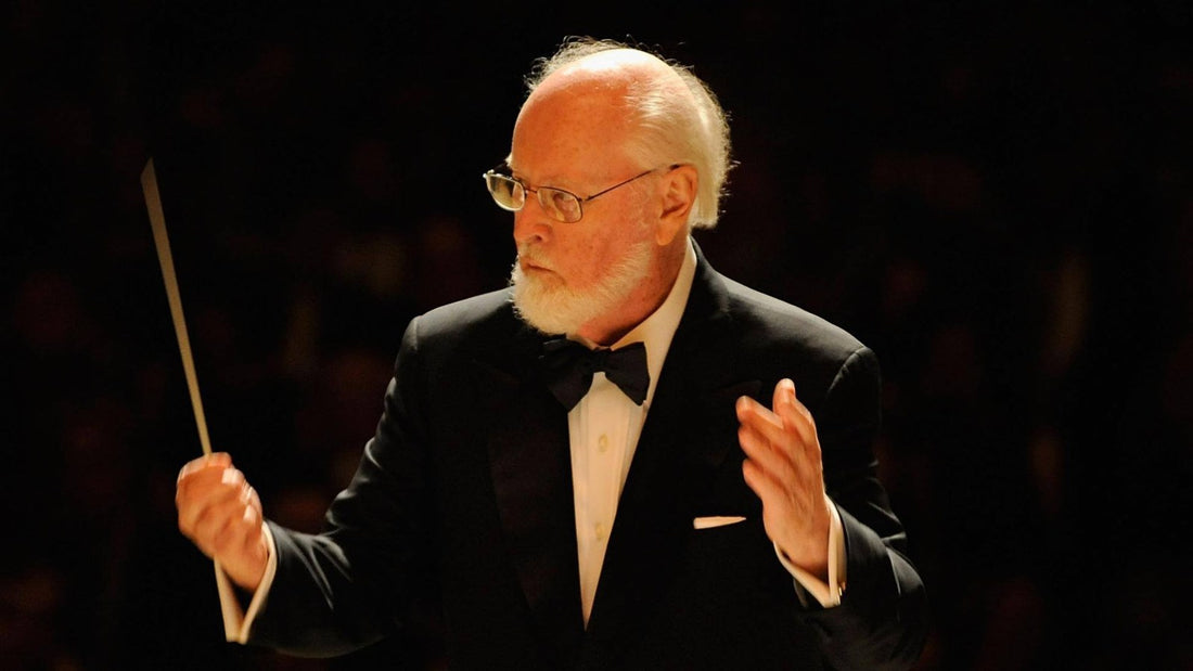 John Williams de smoking, gafas y barba blanca, batuta alzada, dirige una orquesta