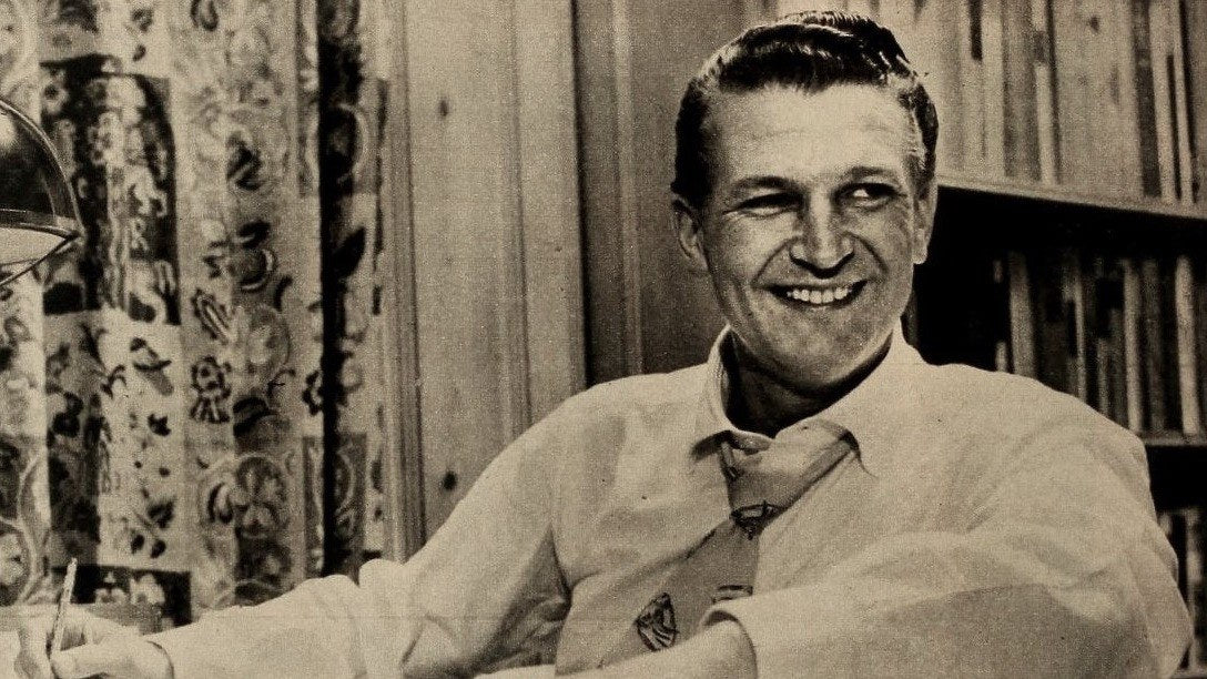 Bud Collyer, la voz de Superman en el serial radiofónico, sonriendo en su escritorio en una foto de 1953