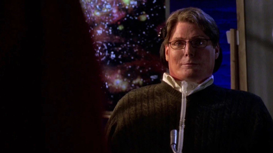 Christopher Reeve como Virgil Swann, con gafas y jersey oscuro, delante de una imagen de estrellas en la serie Smallville